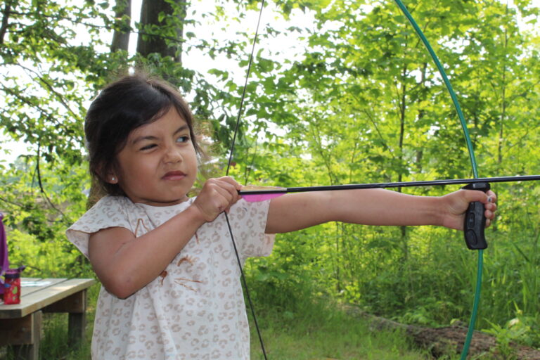 Focused young girl pulling back the string of a green bow at the outdoor archery range.