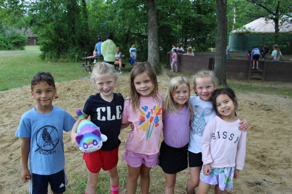 Six young campers smiling and posing together in a sandy play area at summer camp.