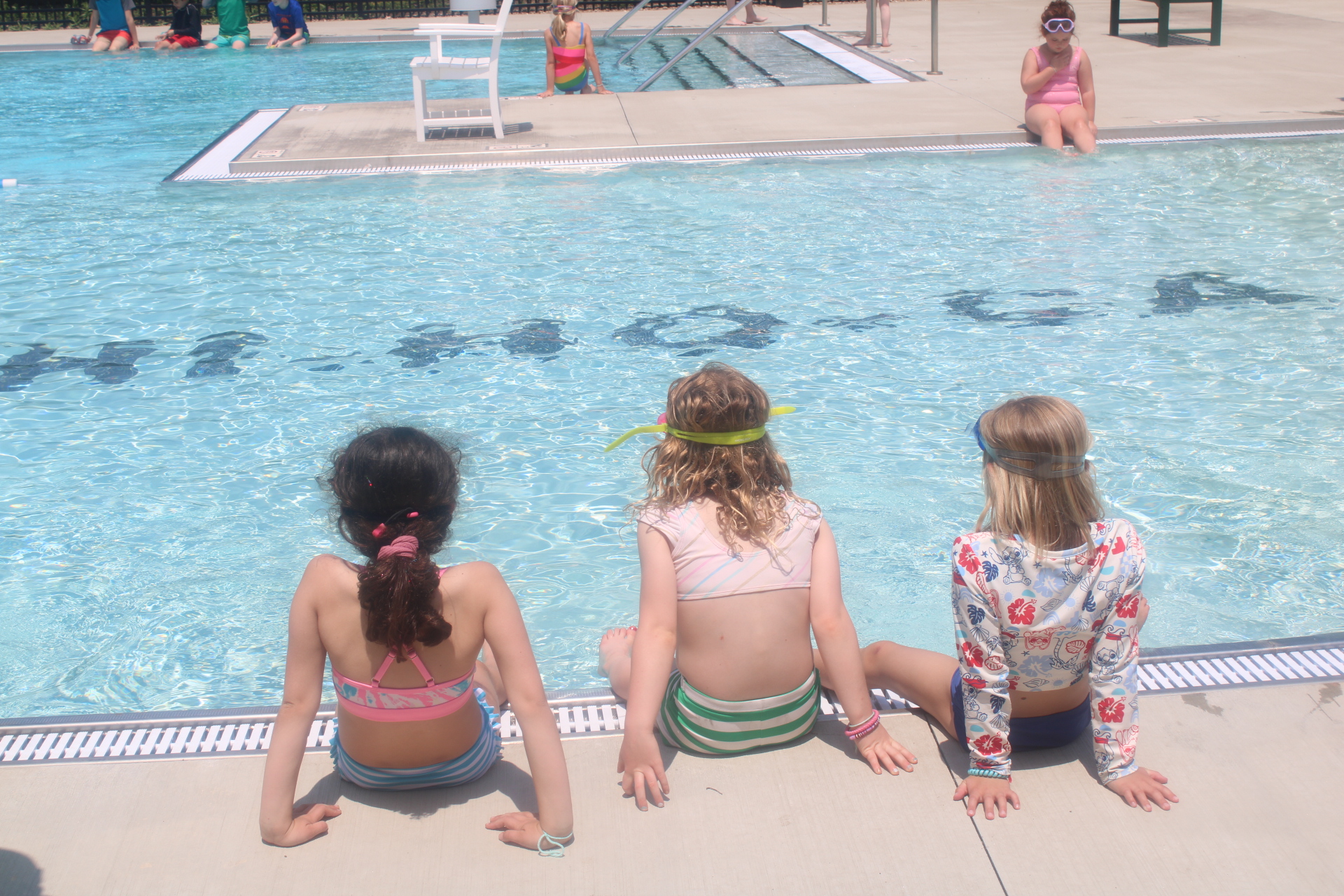 Three girls sitting on the pool deck, dangling their feet into the bright blue swimming pool water.