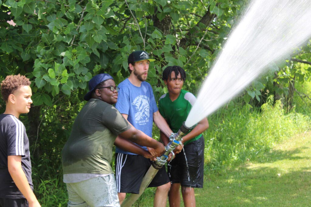 A camp counselor helping two campers hold and spray a large blast of water from a fire hose in a grassy field.