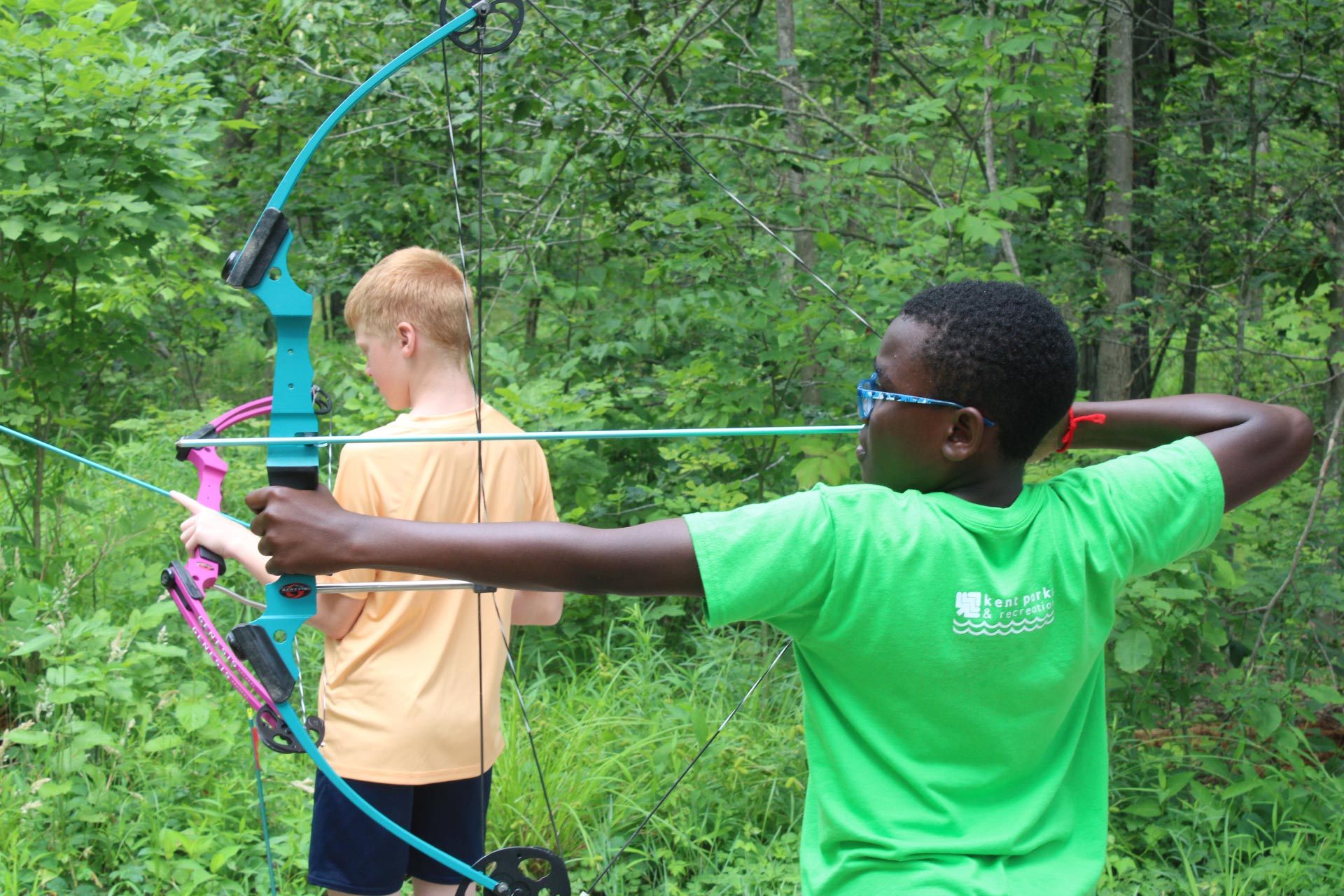 Two young campers drawing back their bows at the outdoor archery range in the woods.