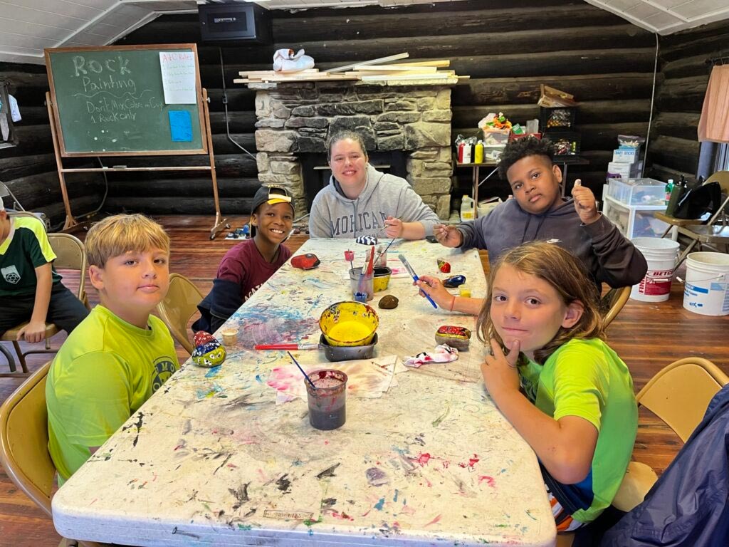 Group of campers and a counselor sitting around a paint-covered table painting small rocks inside a log cabin.
