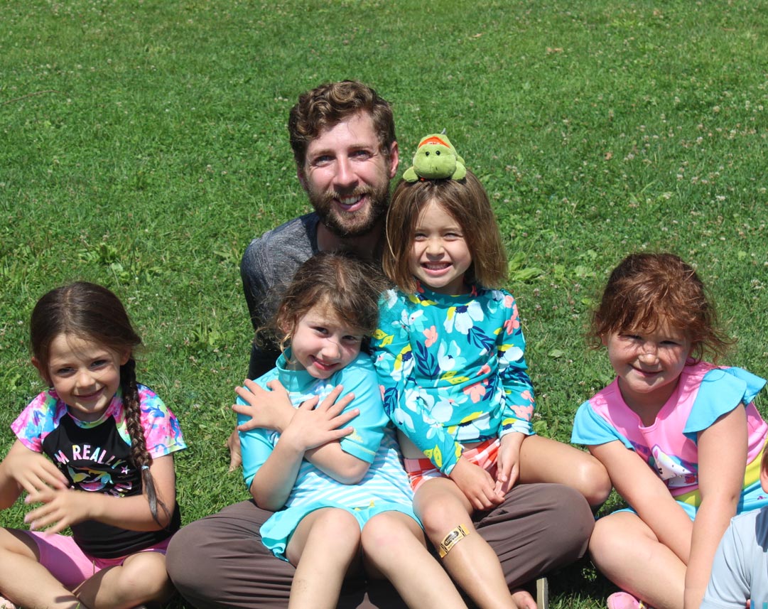 Smiling camp counselor balancing a plush frog toy on a young camper's head while sitting in the grass with a group of kids.