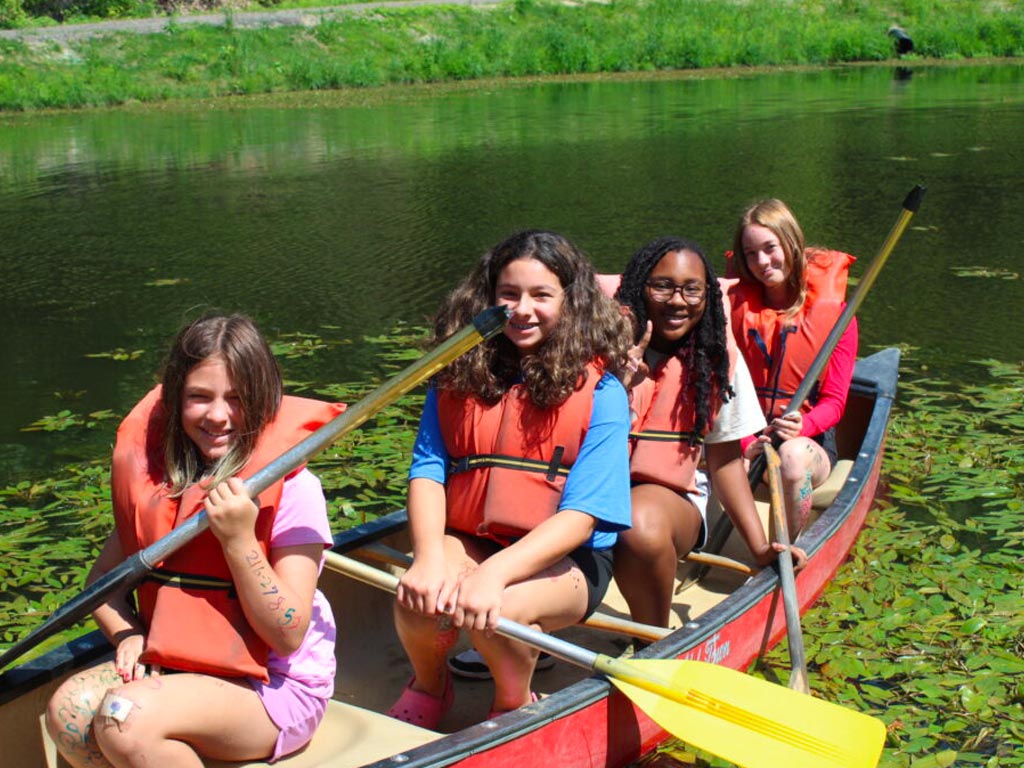 Four smiling campers wearing orange life jackets sitting together in a red canoe on a camp pond.