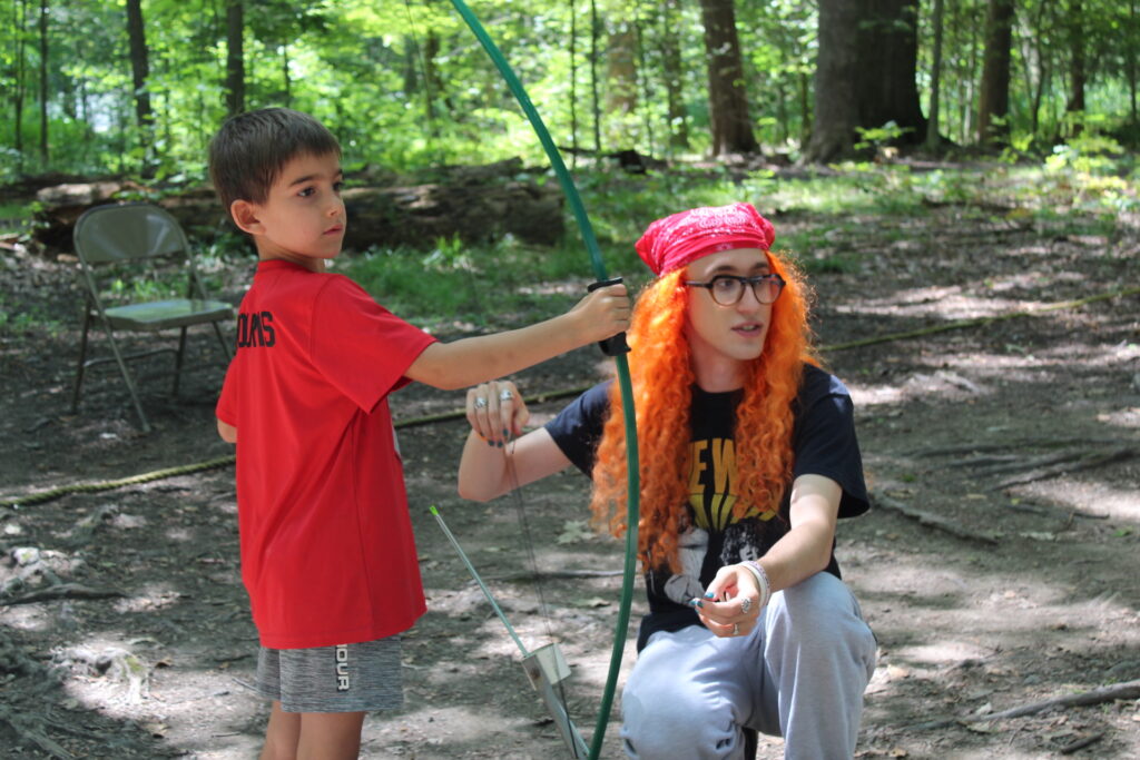 Camp counselor in a fun costume teaching a young camper how to hold a bow and arrow.