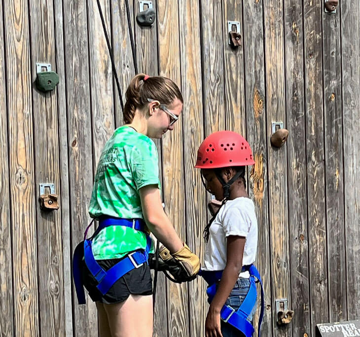 amp staff member securing a red safety helmet and harness on a young camper at the outdoor climbing wall.