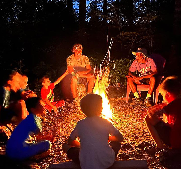 Campers and counselors sitting around a bright campfire at dusk.