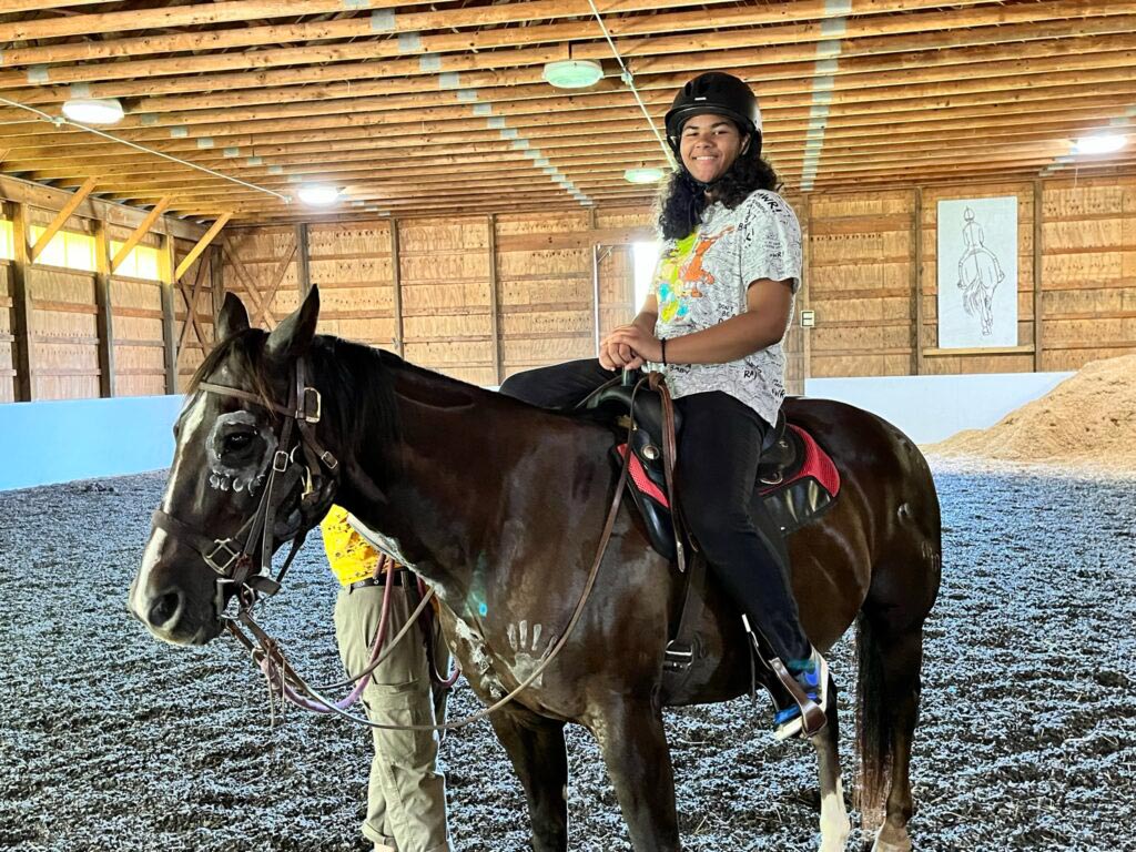 Smiling camper wearing a safety helmet riding a brown horse inside a large indoor equestrian riding arena.