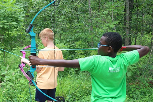 Two young campers drawing back their bows at the outdoor archery range in the woods.