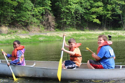 Three smiling campers wearing orange life jackets paddling a silver canoe on a camp pond.
