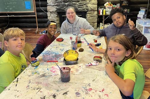 Five smiling campers and a counselor sitting around a large table painting stones inside a rustic log cabin.