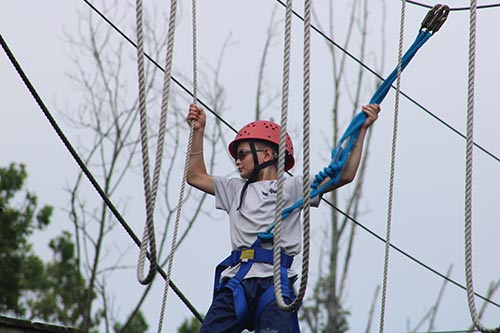 Camper wearing a red helmet and safety harness climbing across elevated ropes in the forest.