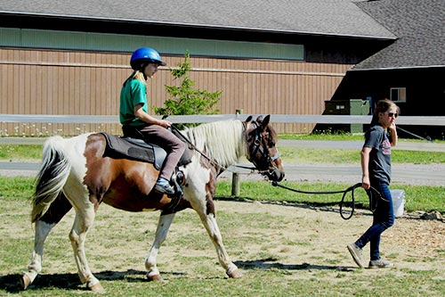 A young camper wearing a blue helmet riding a brown and white horse, being led by a camp staff member.