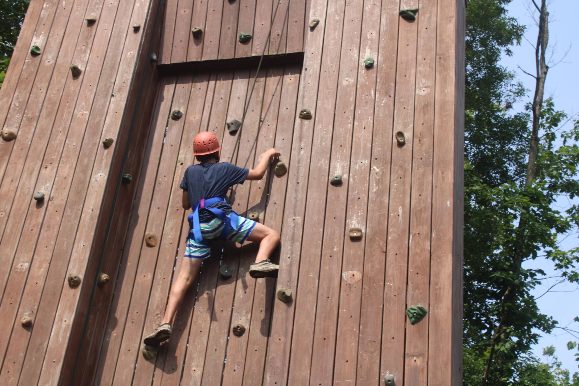 A camper wearing a red helmet and safety harness climbing up a large wooden outdoor climbing wall.