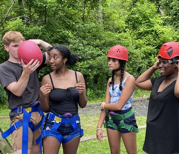 Group of teenagers wearing safety harnesses and putting on red helmets before an adventure activity.