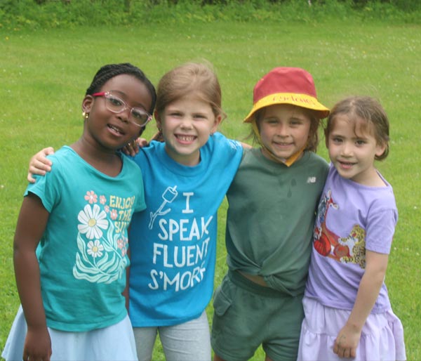 Four young girls standing with their arms around each other smiling in a grassy green field.