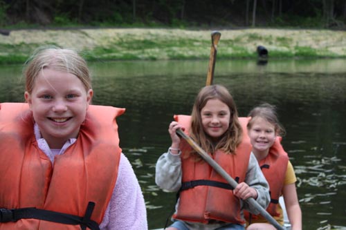 Three smiling girls wearing orange life jackets sitting in a canoe on the water, holding a paddle.