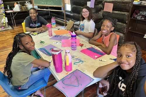 Four young girls sitting at a messy craft table inside a log cabin, smiling with their string art projects.
